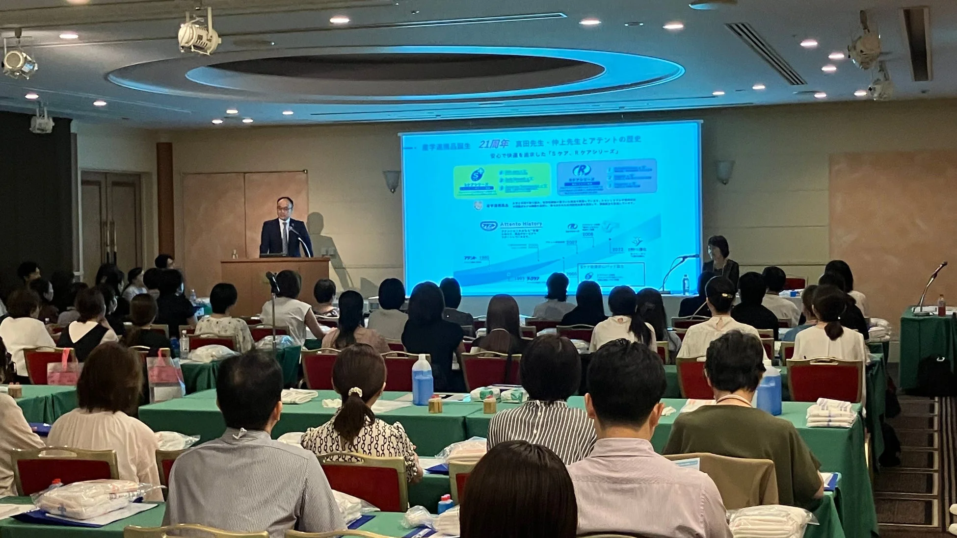 会議室で講演者が壇上からプレゼンテーションを行っており、多くの聴衆が聴講している