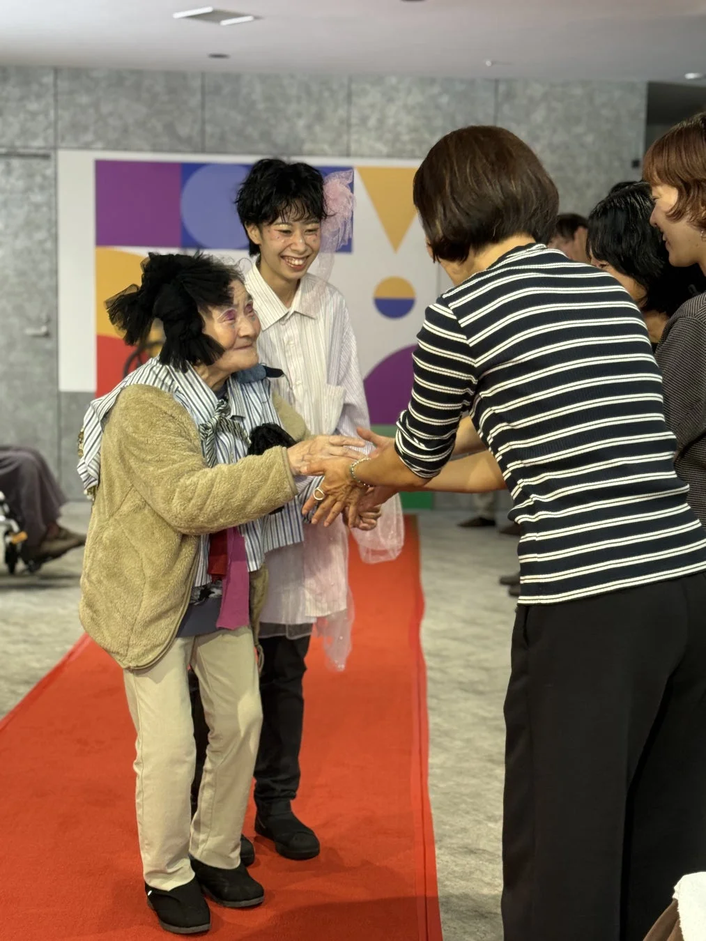 個性的なメイクとヘアスタイルの高齢女性が、レッドカーペットの上で笑顔で交流している様子。若い女性も隣で微笑んでおり、華やかで楽しいイベントの雰囲気が伝わります。