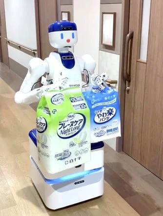 介護施設のような廊下で、白いロボットが大人用紙おむつを運んでいます