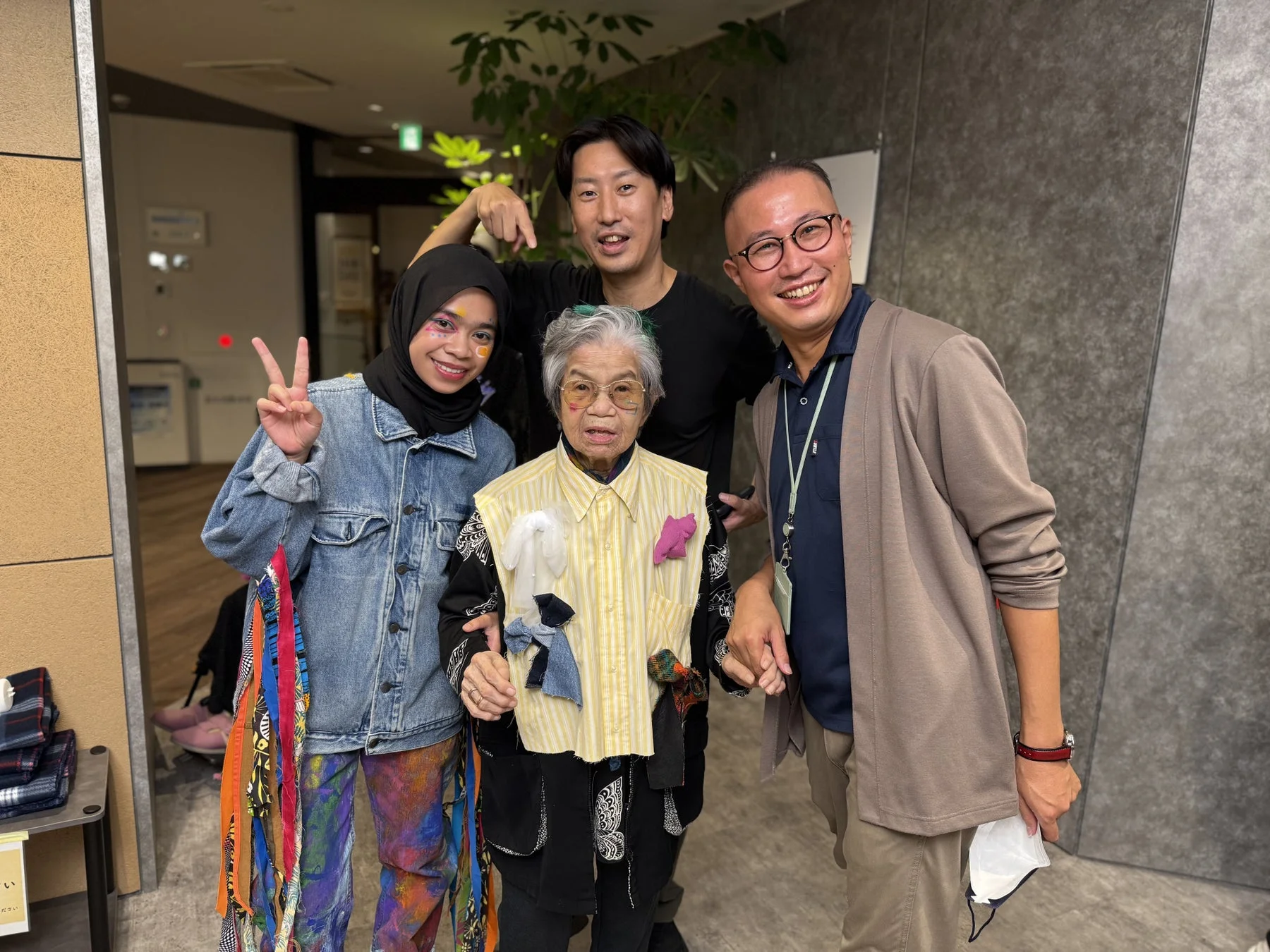 笑顔の男女4人が写る集合写真。ヒジャブの若い女性と、個性的なベストを着た高齢の女性、2人の男性が親しげにポーズをとっている。楽しげな雰囲気のイベントの一コマのようだ。