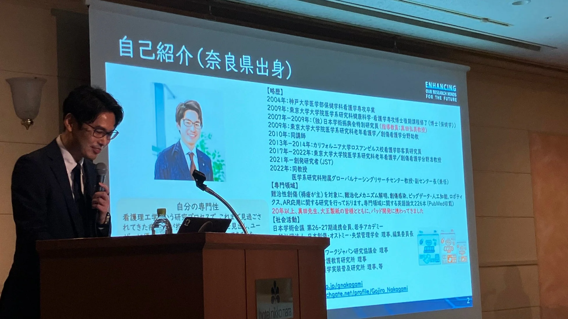 男性が「自己紹介(奈良県出身)」と題されたスライドを背景に講演している