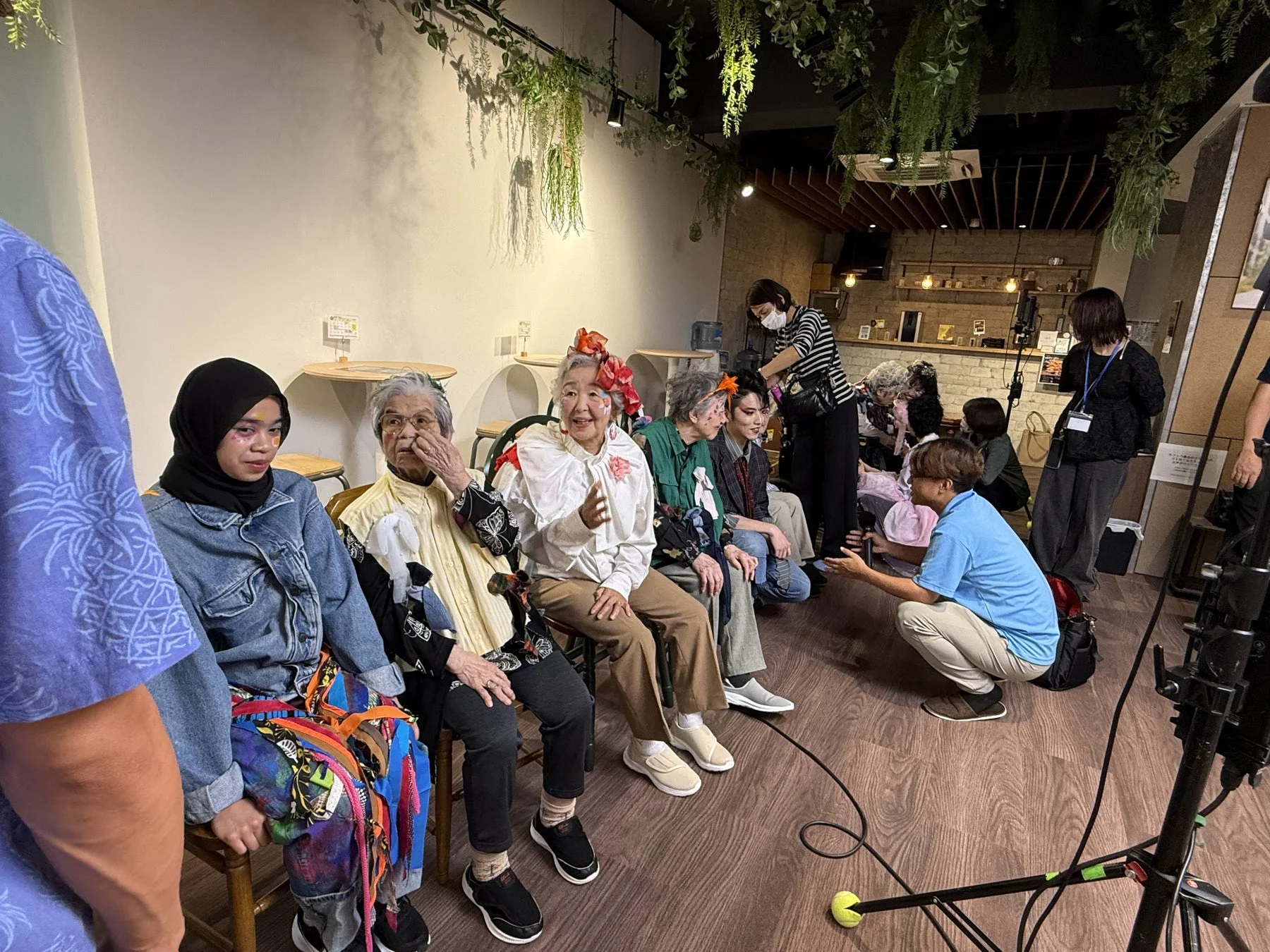 カフェのような空間で、個性的なメイクや服装をした高齢者たちが撮影に参加している様子です。若いスタッフとの交流も見られ、和やかな雰囲気の中で特別なイベントが行われていることが伺えます。