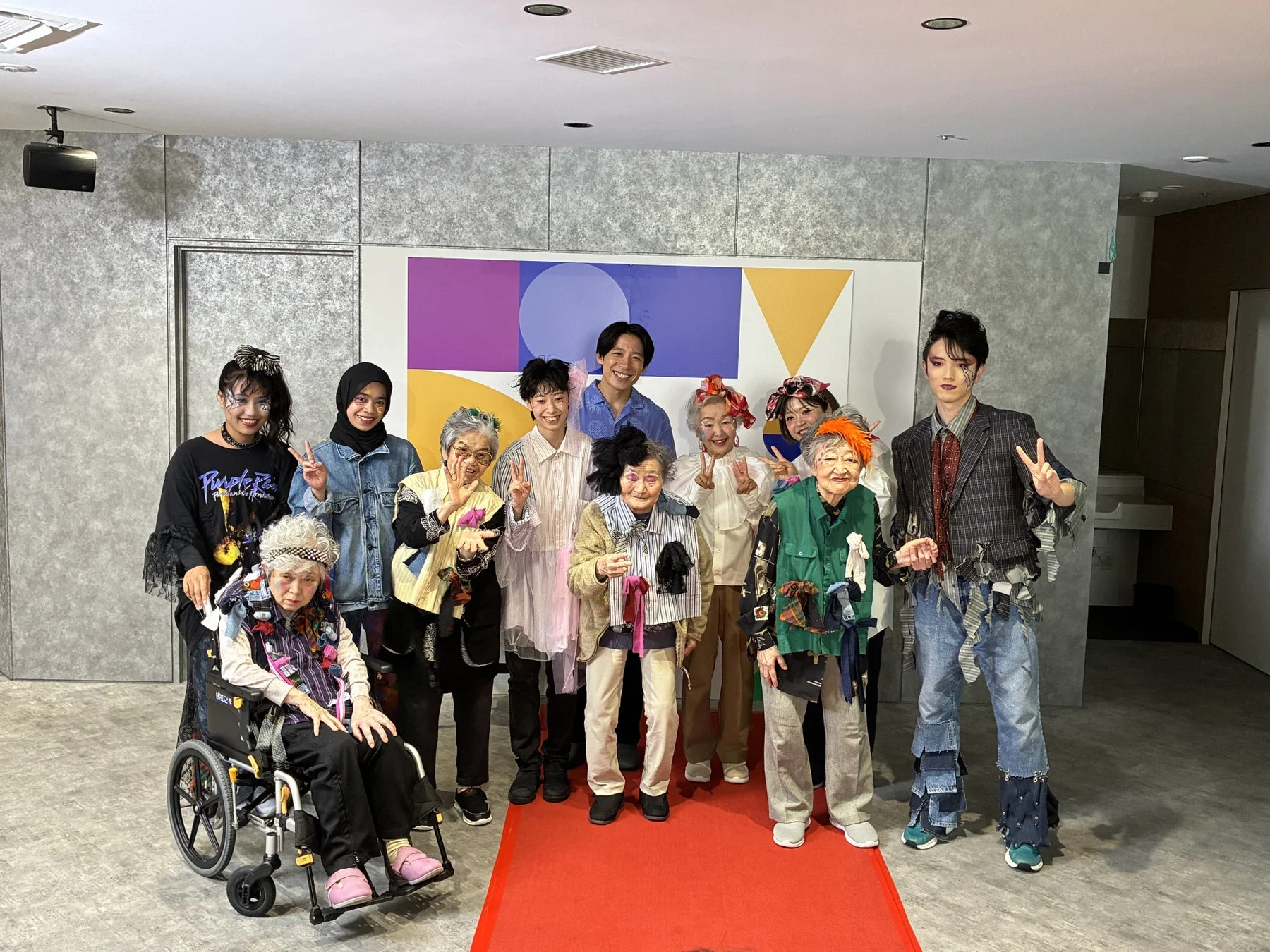 個性的な衣装を身につけた高齢者と若者たちが、レッドカーペットの上で笑顔でポーズをとっている集合写真です。アートイベントかファッションショーの一コマのようです。
